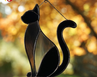 May include: A black cat-shaped stained glass suncatcher. The cat is in a sitting position with a curved tail. The glass has a mix of black and amber tones. It hangs from a chain, perfect for a window or wall decoration.