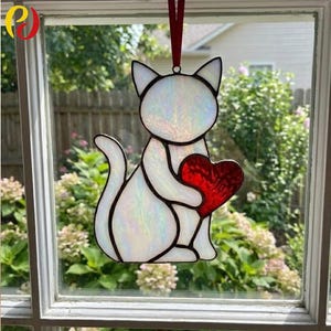 May include: A stained glass cat ornament holding a red heart, suspended by a red ribbon. The cat features white glass with black outlines, and the heart is a bright red. The window art is set against a garden backdrop.