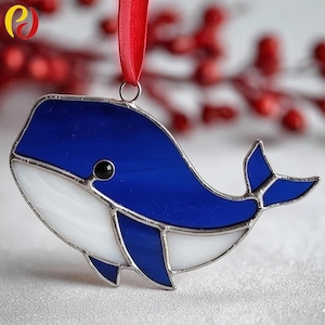 May include: A stained glass whale ornament with a blue body and white belly, accented with a black eye and silver trim. A red ribbon is attached for hanging. The ornament is a decorative item.