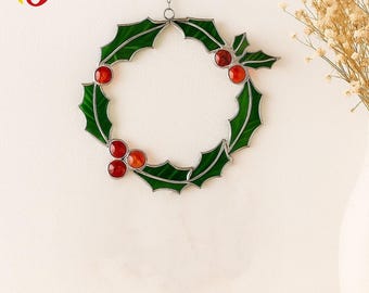 Op de afbeelding: Een gebrandschilderde kerstkrans met groene hulstbladeren en rode bessen. De cirkelvormige krans hangt aan een zilveren ketting. Het ontwerp is geplaatst tegen een witte achtergrond.