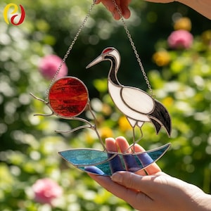 May include: Stained glass crane suncatcher with a red sun, white and black crane, and blue water design. The suncatcher is suspended by a silver chain. The background is a blurred garden scene with pink flowers.