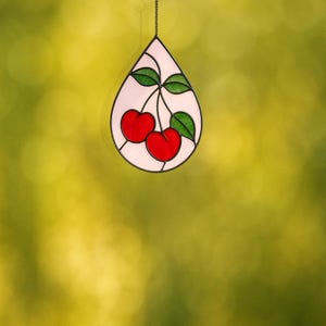 Może przedstawiać: Wiszący witraż w kształcie łzy, przedstawiający dwie czerwone wiśnie z zielonymi liśćmi. Okno jest obrysowane na czarno i zawieszone na delikatnym łańcuszku. Tło jest rozmyte, żółto-zielone.