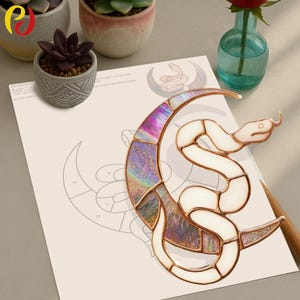 Op de afbeelding: Een glas-in-lood ontwerp van een slang en een halve maan met iriserend en wit glas, koperkleurig metaal en een gedetailleerd patroon. Het kunstwerk is geplaatst op een papier met een tekening van het ontwerp. Het thema is mystiek en hemels.