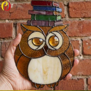 Puede incluir: Figura de búho de vidriera con una pila de libros en la cabeza. El búho presenta vidrio marrón, ámbar y crema, con un diseño detallado. Los libros son rojos, azules, verdes y morados. Un artículo decorativo para los amantes de los libros.