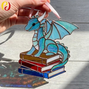Peut inclure: Ornement en vitrail représentant un dragon bleu et turquoise, perché sur une pile de livres. Le dragon a des cornes et des ailes blanches, et un ruban rose pour l'accrocher. Les livres sont marron, blancs, rouges et bleus.