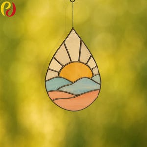 May include: A stained glass suncatcher in a teardrop shape. The design features a yellow sun, blue and orange landscape, and white sky. The suncatcher hangs from a chain and is set against a blurred green background.