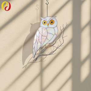Könnte beinhalten: Ein Glas-Eule-Suncatcher mit weißem Körper, schillernden Flügeln und gelben Augen. Die Eule sitzt auf einem Metallast und hängt an einer Kette. Der Suncatcher wirft einen Schatten an die Wand.