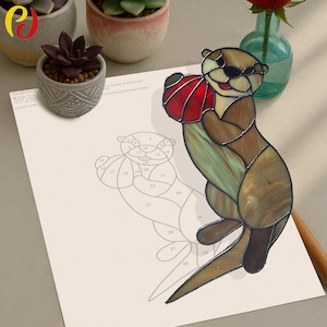 Pode incluir: Uma estatueta de lontra em vitral segurando uma bola vermelha. A lontra tem painéis de vidro marrom, verde e bronzeado. Um desenho a traço da lontra está em papel branco. A lontra é um objeto decorativo.