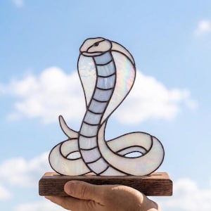May include: A stained glass cobra sculpture featuring iridescent white glass panels with dark solder lines. The snake is coiled in an upright hood display and is mounted on a rectangular wooden base, showcasing a reflective surface texture.