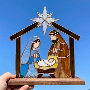 May include: A stained glass nativity scene featuring a central star with iridescent glass, a blue-robed figure, a brown-robed figure, and a baby in a yellow manger, all framed by a dark brown structure mounted on a rectangular wooden base.