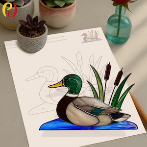 May include: A stained glass duck with green head, brown body, and yellow beak, resting on a blue base. The duck is surrounded by green reeds and cattails. A drawing of a duck is on the paper behind the glass duck.