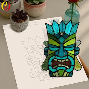 Op de afbeelding: Een kleurrijk tiki-masker van glas in lood met tinten blauw, groen en bruin. Het masker ligt op een vel papier met een genummerd diagram. Het masker heeft een gedetailleerd ontwerp met een kroon en open mond.