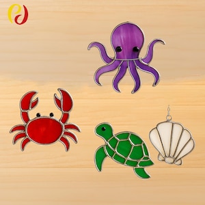Puede incluir: Cuatro criaturas marinas de vidrieras: un cangrejo rojo, un pulpo morado, una tortuga verde y una concha blanca. Cada pieza tiene un borde plateado y está sobre un fondo de madera clara.