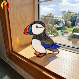 Puede incluir: Figura de frailecillo de vidriera. El ave tiene cabeza negra, pecho blanco, alas grises con detalles azules y pico colorido. La figura se exhibe en un alféizar de madera.