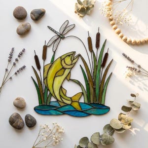 Peut inclure: Un attrape-soleil en vitrail représentant un poisson jaune sautant hors de l'eau bleue, entouré de roseaux verts et d'une libellule. La pièce est assemblée avec des lignes de soudure en métal sombre, formant une scène naturelle aux couleurs vives.