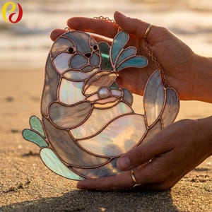 Pode incluir: Apanhador de sol de vidro manchado de uma lontra com vidro iridescente e folha de cobre. A lontra está segurando uma pérola e tem detalhes em azul e verde. O apanhador de sol está pendurado em uma corrente.