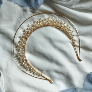 May include: A gold-colored metal headband with a half-circle design, adorned with numerous small, white pearls. The pearls are clustered along the top of the headband, creating a crown-like appearance. The headband is set against a white fabric background.