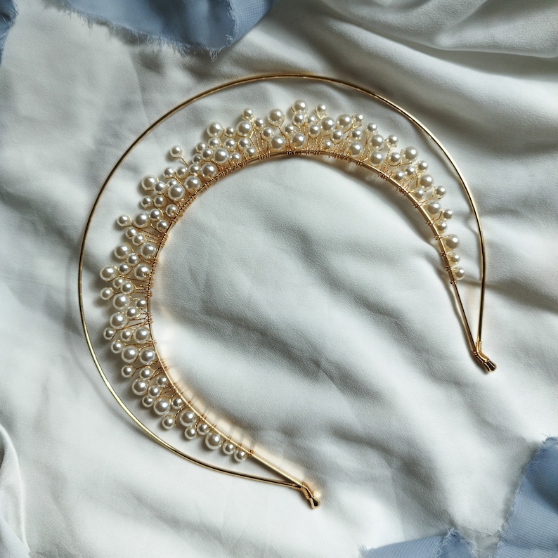 May include: A gold-colored metal headband with a half-circle design, adorned with numerous small, white pearls. The pearls are clustered along the top of the headband, creating a crown-like appearance. The headband is set against a white fabric background.