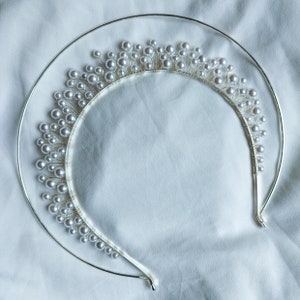 May include: A silver-toned bridal tiara with a halo design. The tiara features a series of small, white, pearl-like beads attached to thin silver wires, creating a delicate, elegant appearance. The tiara is set against a white fabric background.