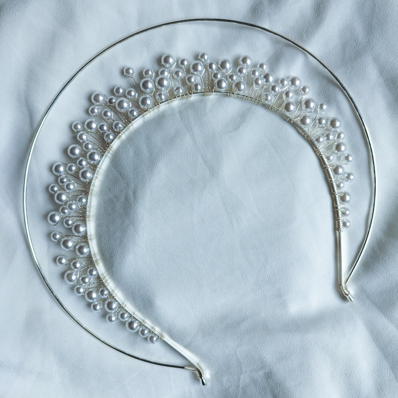 May include: A silver-toned bridal tiara with a halo design. The tiara features a series of small, white, pearl-like beads attached to thin silver wires, creating a delicate, elegant appearance. The tiara is set against a white fabric background.