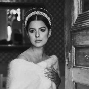 May include: A black and white portrait of a person wearing a halo-style headpiece adorned with pearls and crystals. The person is also wearing a white fur stole. The background includes a wooden door and a patterned wall.