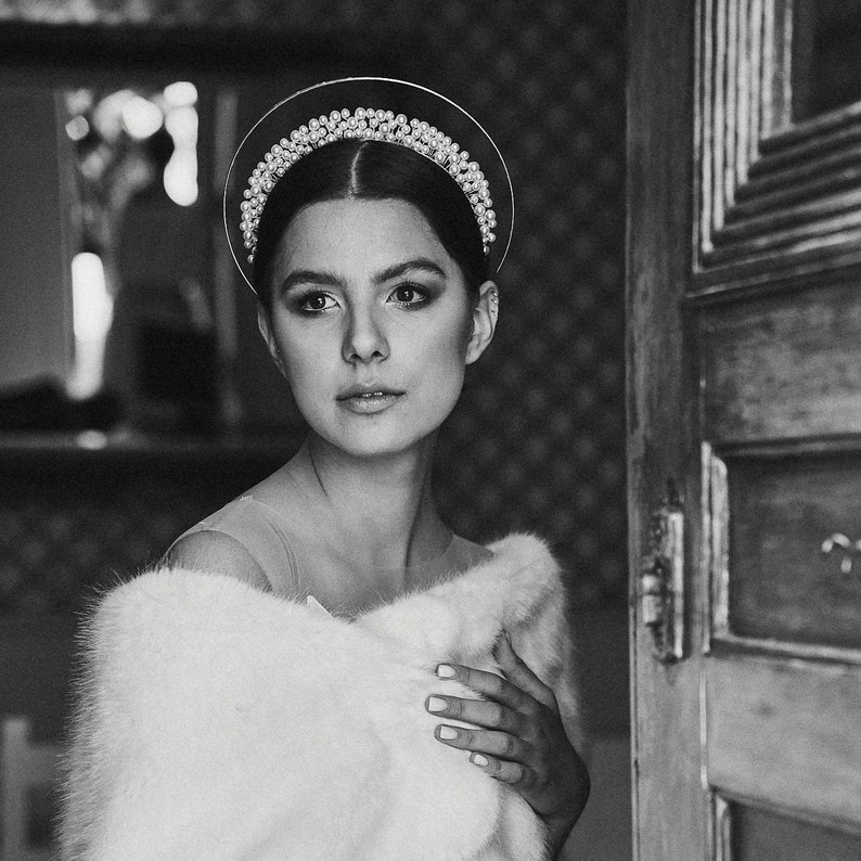 May include: A black and white portrait of a person wearing a halo-style headpiece adorned with pearls and crystals. The person is also wearing a white fur stole. The background includes a wooden door and a patterned wall.