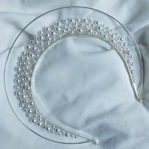 May include: A silver-toned tiara with a delicate design. The tiara features a series of clustered, round, white pearls. The pearls are attached to a thin, curved metal band. The tiara is set against a white, textured background.