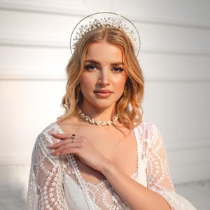 May include: A woman wearing a pearl necklace, earrings, and a ring. She is also wearing a white lace dress and a halo-style headpiece adorned with pearls. The woman has blonde hair and is looking at the camera.