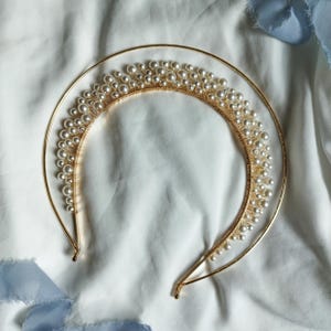 May include: A gold-toned bridal headband featuring a crescent-shaped design adorned with numerous faux pearls. The headband has a delicate, elegant appearance, suitable for weddings or special occasions. The pearls are clustered along the top of the headband.