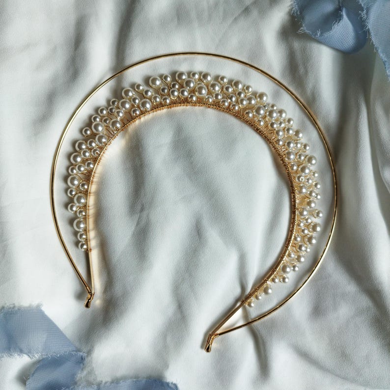 May include: A gold-toned bridal headband featuring a crescent-shaped design adorned with numerous faux pearls. The headband has a delicate, elegant appearance, suitable for weddings or special occasions. The pearls are clustered along the top of the headband.