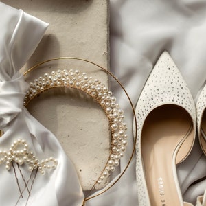 May include: A bridal ensemble featuring a gold-toned pearl-studded tiara, matching hairpins, and white pointed-toe heels adorned with rhinestones. The items rest on a white satin fabric, creating a luxurious and elegant aesthetic. The tiara has a circular design.