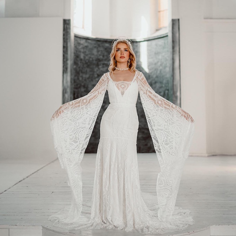 May include: A white lace wedding dress with long, flowing sleeves and a fitted bodice. The dress features a V-neckline and a mermaid silhouette. The bride is wearing a beaded headband and necklace. The dress is displayed in a bright, open space.