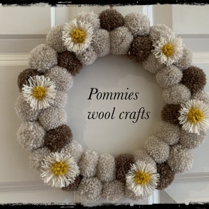 K&ouml;nnte beinhalten: Ein Kranz aus braunen und beigefarbenen Pompons mit wei&szlig;en, g&auml;nsebl&uuml;mchenartigen Bl&uuml;ten. Der Kranz tr&auml;gt den Text "Pommies wool crafts" in der Mitte.