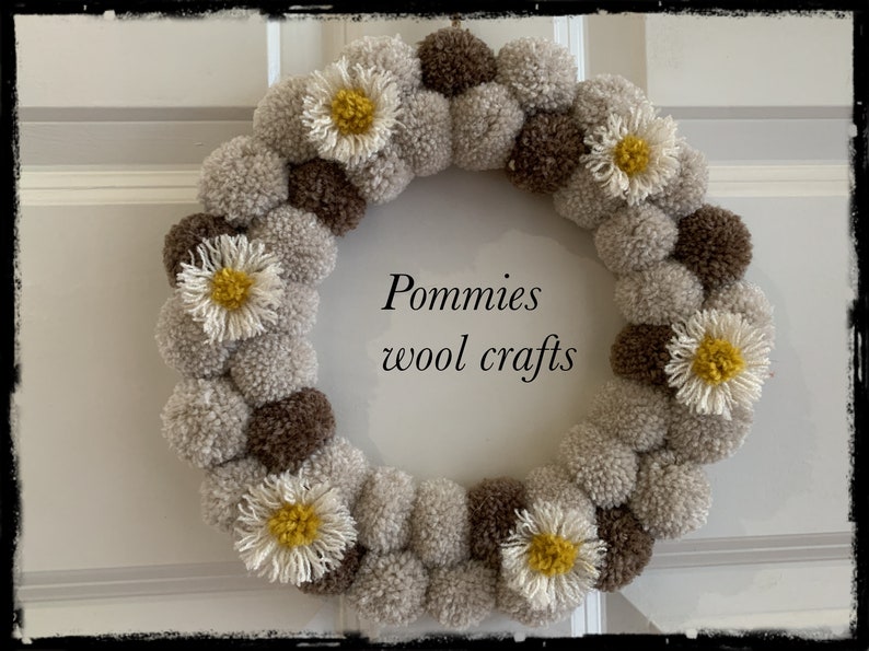 K&ouml;nnte beinhalten: Ein Kranz aus braunen und beigefarbenen Pompons mit wei&szlig;en, g&auml;nsebl&uuml;mchenartigen Bl&uuml;ten. Der Kranz tr&auml;gt den Text "Pommies wool crafts" in der Mitte.