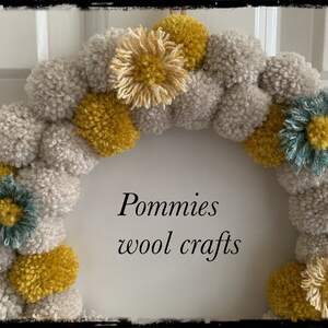 K&ouml;nnte beinhalten: Ein Kranz aus grauen und gelben Pompons mit blauen und gelben Blumenakzenten. Der Kranz tr&auml;gt den Text "Pommies wool crafts" in der Mitte.