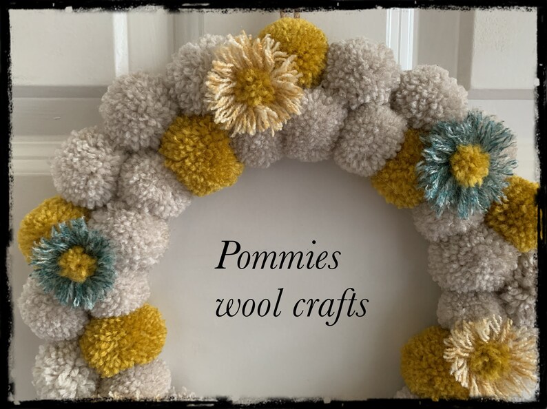 K&ouml;nnte beinhalten: Ein Kranz aus grauen und gelben Pompons mit blauen und gelben Blumenakzenten. Der Kranz tr&auml;gt den Text "Pommies wool crafts" in der Mitte.