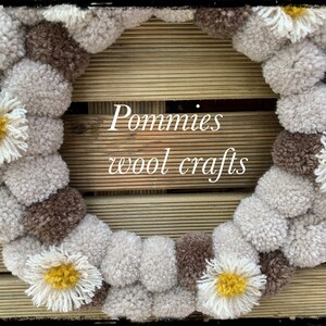 K&ouml;nnte beinhalten: Ein Kranz aus braunen und wei&szlig;en Pompons mit gelben und wei&szlig;en G&auml;nsebl&uuml;mchen-Akzenten. Der Kranz befindet sich auf einem Holzgrund mit dem Text "Pommies wool crafts".
