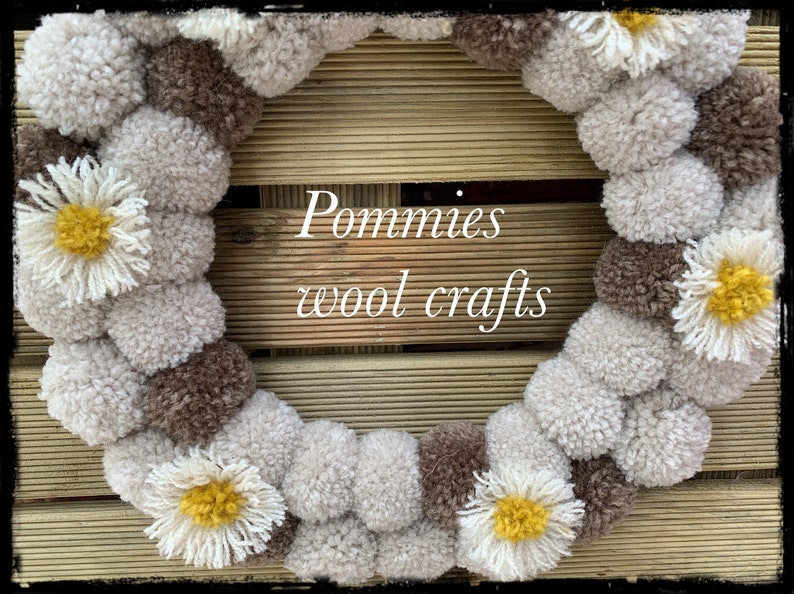 K&ouml;nnte beinhalten: Ein Kranz aus braunen und wei&szlig;en Pompons mit gelben und wei&szlig;en G&auml;nsebl&uuml;mchen-Akzenten. Der Kranz befindet sich auf einem Holzgrund mit dem Text "Pommies wool crafts".