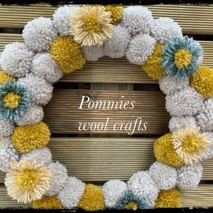 K&ouml;nnte beinhalten: Ein Kranz aus wei&szlig;en, gelben und t&uuml;rkisfarbenen Pompons. Der Kranz ist mit verschiedenen Pompons in unterschiedlichen Gr&ouml;&szlig;en und Farben verziert. Der Kranz befindet sich auf einem Holzgrund. Der Text "Pommies wool crafts" befindet sich in der Mitte des Kranzes.