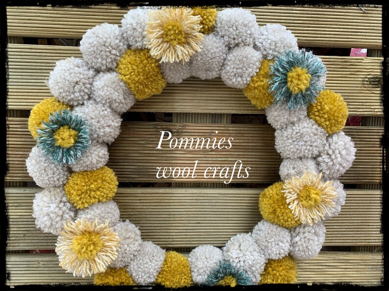 K&ouml;nnte beinhalten: Ein Kranz aus wei&szlig;en, gelben und t&uuml;rkisfarbenen Pompons. Der Kranz ist mit verschiedenen Pompons in unterschiedlichen Gr&ouml;&szlig;en und Farben verziert. Der Kranz befindet sich auf einem Holzgrund. Der Text "Pommies wool crafts" befindet sich in der Mitte des Kranzes.