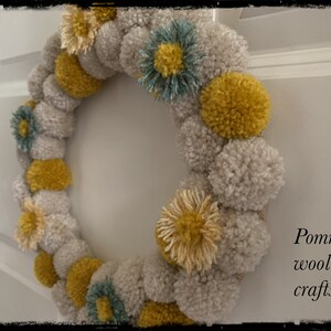 K&ouml;nnte beinhalten: Ein Kranz aus beige und gelben Pompons mit einigen blauen Pompons. Der Kranz ist mit kleinen gelben und beigen Blumen verziert. Der Text "Pommies wool crafts" ist in der rechten unteren Ecke des Bildes zu sehen.