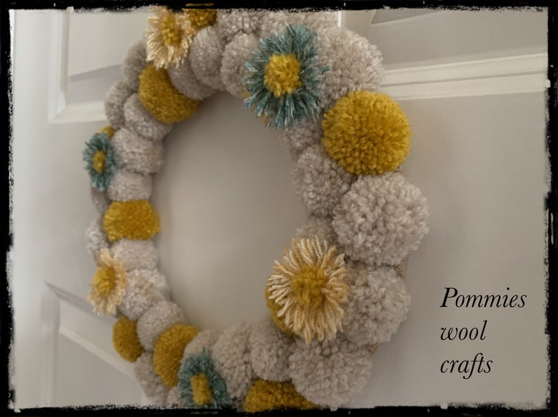 K&ouml;nnte beinhalten: Ein Kranz aus beige und gelben Pompons mit einigen blauen Pompons. Der Kranz ist mit kleinen gelben und beigen Blumen verziert. Der Text "Pommies wool crafts" ist in der rechten unteren Ecke des Bildes zu sehen.