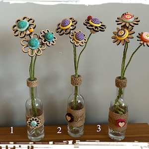 Op de afbeelding: Drie glazen flessen met jute gewikkeld rond de boven- en onderkant, elk met een andere opstelling van houten bloemen met stoffen knoopcentra. De bloemen zijn in tinten blauw, paars, geel en rood.