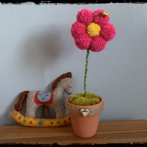 Peut inclure: Une fleur en laine rose avec un centre jaune est placée dans un pot en terre cuite avec un ruban en toile de jute et deux cœurs en bois. La fleur est sur une surface en bois à côté d'un cheval à bascule en feutre gris.