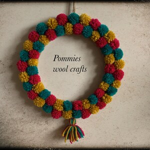 K&ouml;nnte beinhalten: Ein Kranz aus roten, gelben und t&uuml;rkisfarbenen Pompons. Der Kranz h&auml;ngt an einer Wand und hat einen kleinen Quasten, der von unten herabh&auml;ngt. Der Text "Pommies wool crafts" befindet sich in der Mitte des Kranzes.