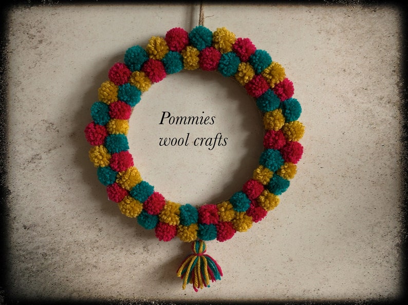 K&ouml;nnte beinhalten: Ein Kranz aus roten, gelben und t&uuml;rkisfarbenen Pompons. Der Kranz h&auml;ngt an einer Wand und hat einen kleinen Quasten, der von unten herabh&auml;ngt. Der Text "Pommies wool crafts" befindet sich in der Mitte des Kranzes.