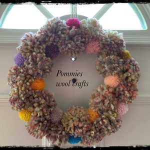 K&ouml;nnte beinhalten: Ein bunter Kranz aus Wollpompons in den Farben Rosa, Lila, Gelb, Orange und Blau. Der Kranz h&auml;ngt an einer wei&szlig;en T&uuml;r mit dem Text "Pommies wool crafts" in Schwarz.