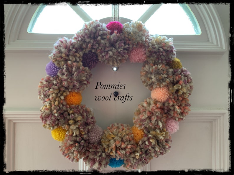 K&ouml;nnte beinhalten: Ein bunter Kranz aus Wollpompons in den Farben Rosa, Lila, Gelb, Orange und Blau. Der Kranz h&auml;ngt an einer wei&szlig;en T&uuml;r mit dem Text "Pommies wool crafts" in Schwarz.