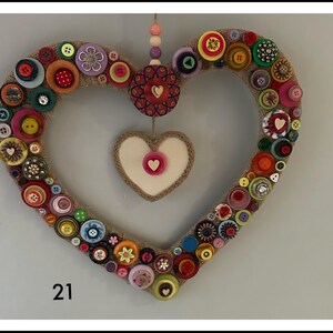 May include: A heart-shaped wall hanging covered in a variety of colourful buttons in shades of red, blue, green, yellow, and purple. A smaller heart with a cream centre and a pink button hangs in the middle. The number 21 is printed in the corner.