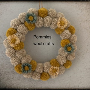 K&ouml;nnte beinhalten: Ein beige-gelber Pompon-Kranz mit kleinen Blumenakzenten. Der Kranz tr&auml;gt den Text "Pommies wool crafts" in der Mitte.